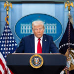 Man speaking at podium with flags in background